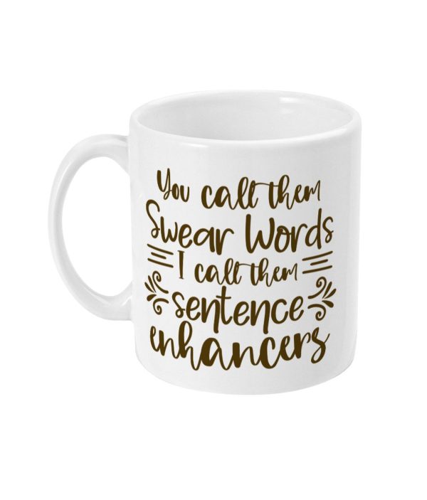 White ceramic mug with golden text 'You call them swear words I call them sentence enhancers' surrounded by decorative swirls and leaf-like motifs.