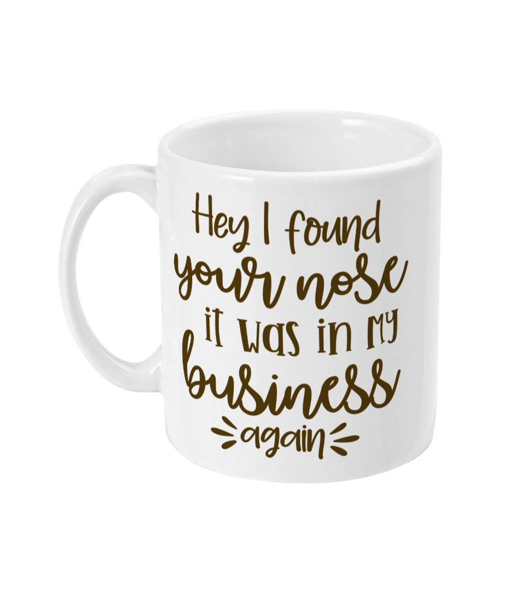 White mug with humorous text 'Hey I found your nose it was in my business again' in decorative brown font and whimsical design elements.